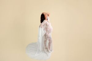 Pregnant woman in elegant white lace maternity gown with flowing cape, posing gracefully against a soft beige background.