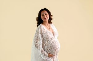 Pregnant woman in white lace dress cradling baby bump, eyes closed, smiling softly against cream background.
