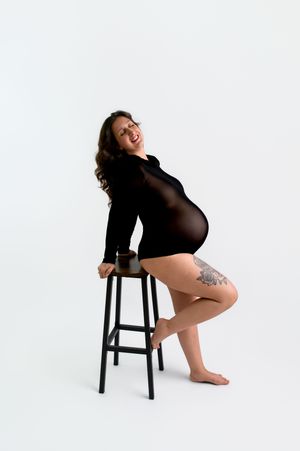 Smiling pregnant woman in black bodysuit sitting on a bar stool, showing baby bump and leg tattoo against white background.