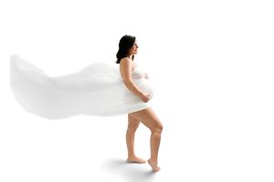 Pregnant woman draped in flowing white fabric poses gracefully against a white background, showcasing her baby bump.
