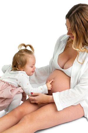 Pregnant woman in white robe sitting with toddler girl touching her baby bump during maternity photo session.