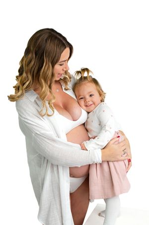 Pregnant woman in white cardigan holds smiling toddler girl in pink skirt during maternity photo session.