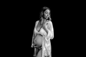 Black and white maternity photo of a pregnant woman in lace robe cradling her baby bump against dark background.