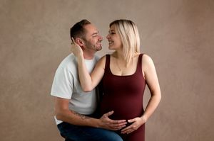 Couple sharing a tender moment during maternity photo shoot, pregnant woman in burgundy dress with partner on brown backdrop.