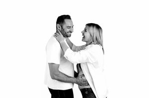 Black and white photo of a smiling couple embracing, woman placing hands on mans neck in a studio setting.