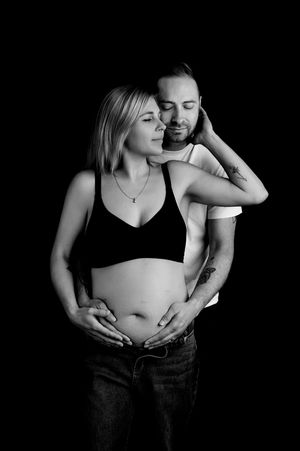 Black and white maternity photo of couple embracing, man kissing womans neck while both cradle her pregnant belly.