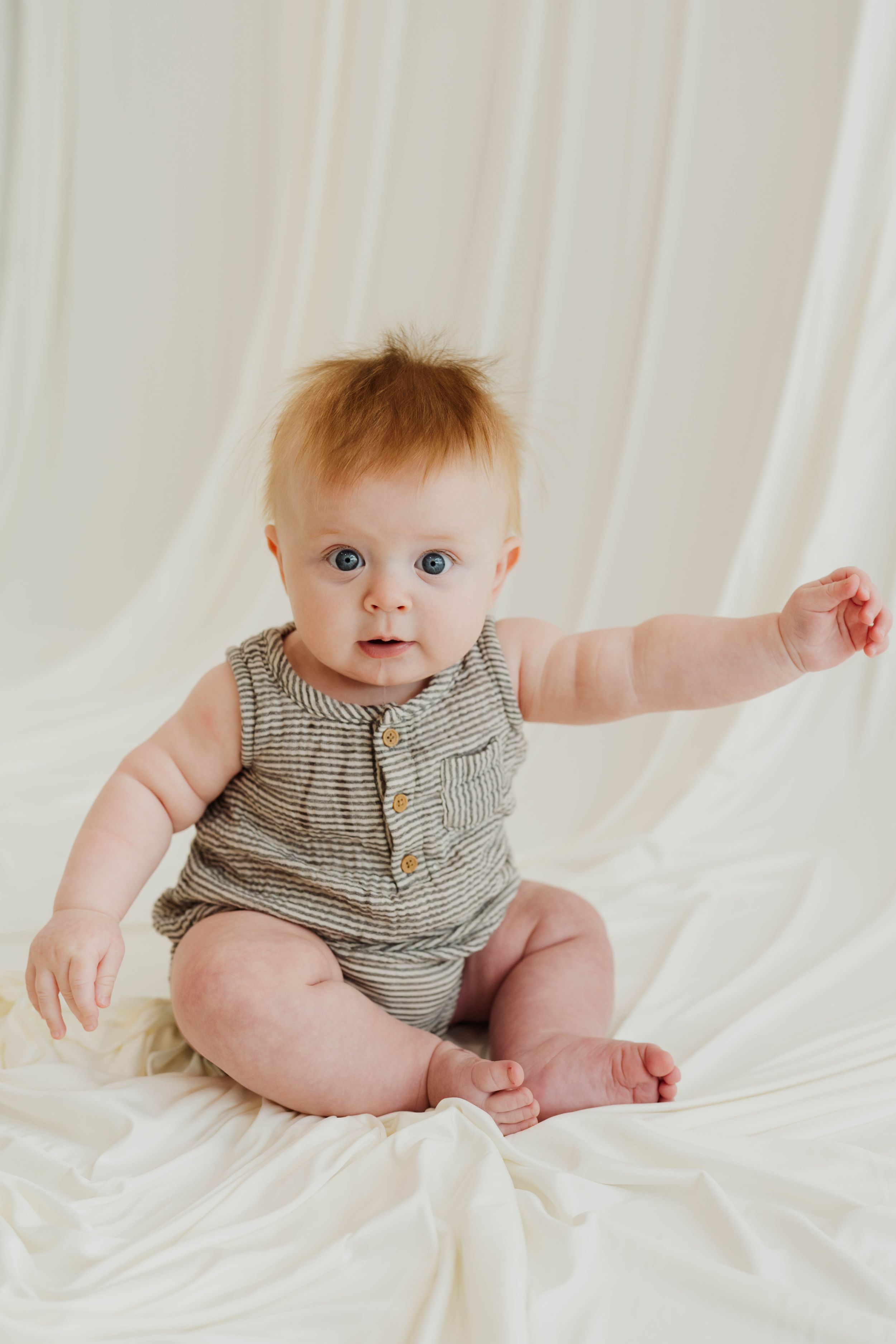Adorable baby with blue eyes and red hair sits on white fabric wearing a striped romper with wooden buttons.