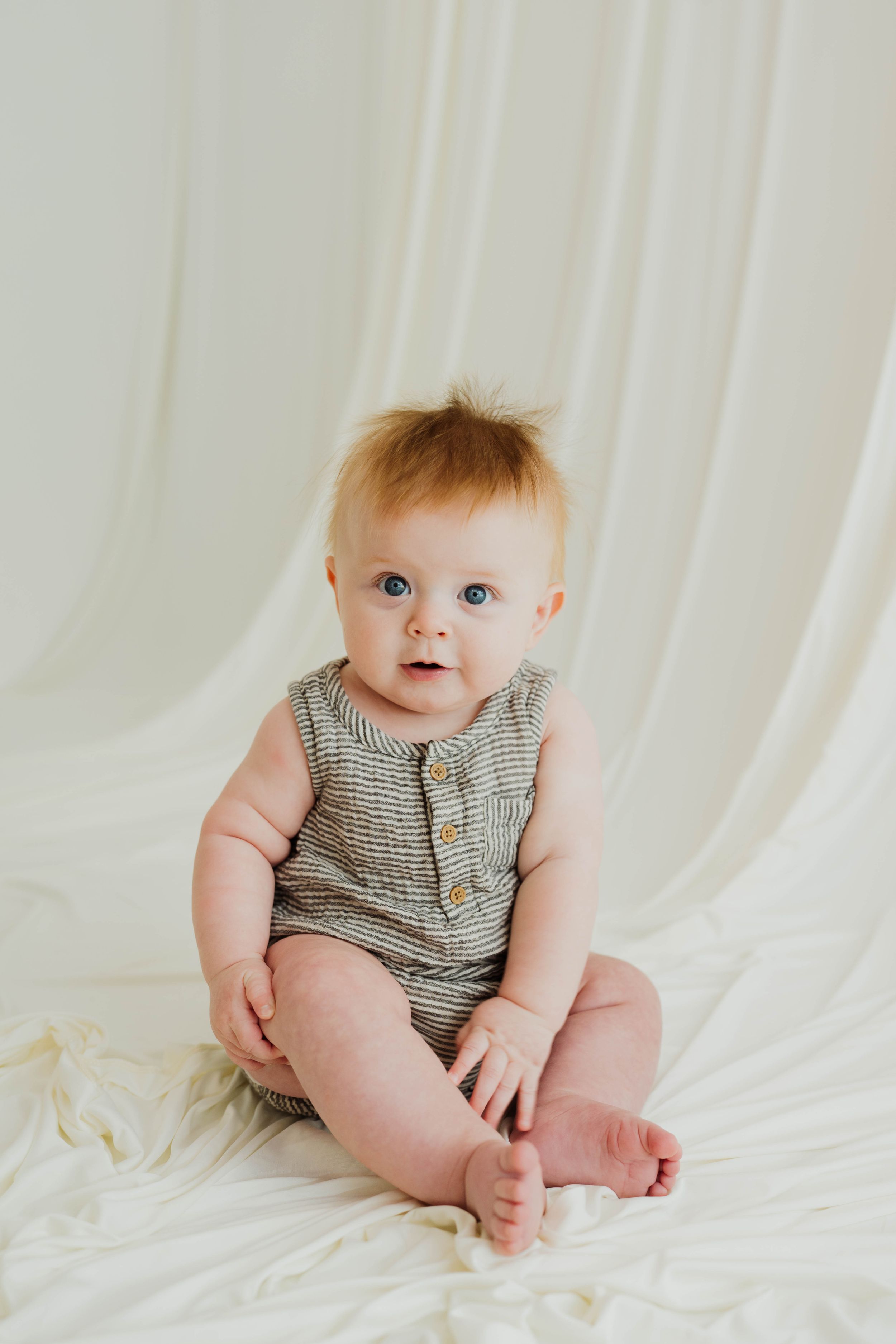 A cute baby with blue eyes sits on a white fabric backdrop wearing a striped romper.