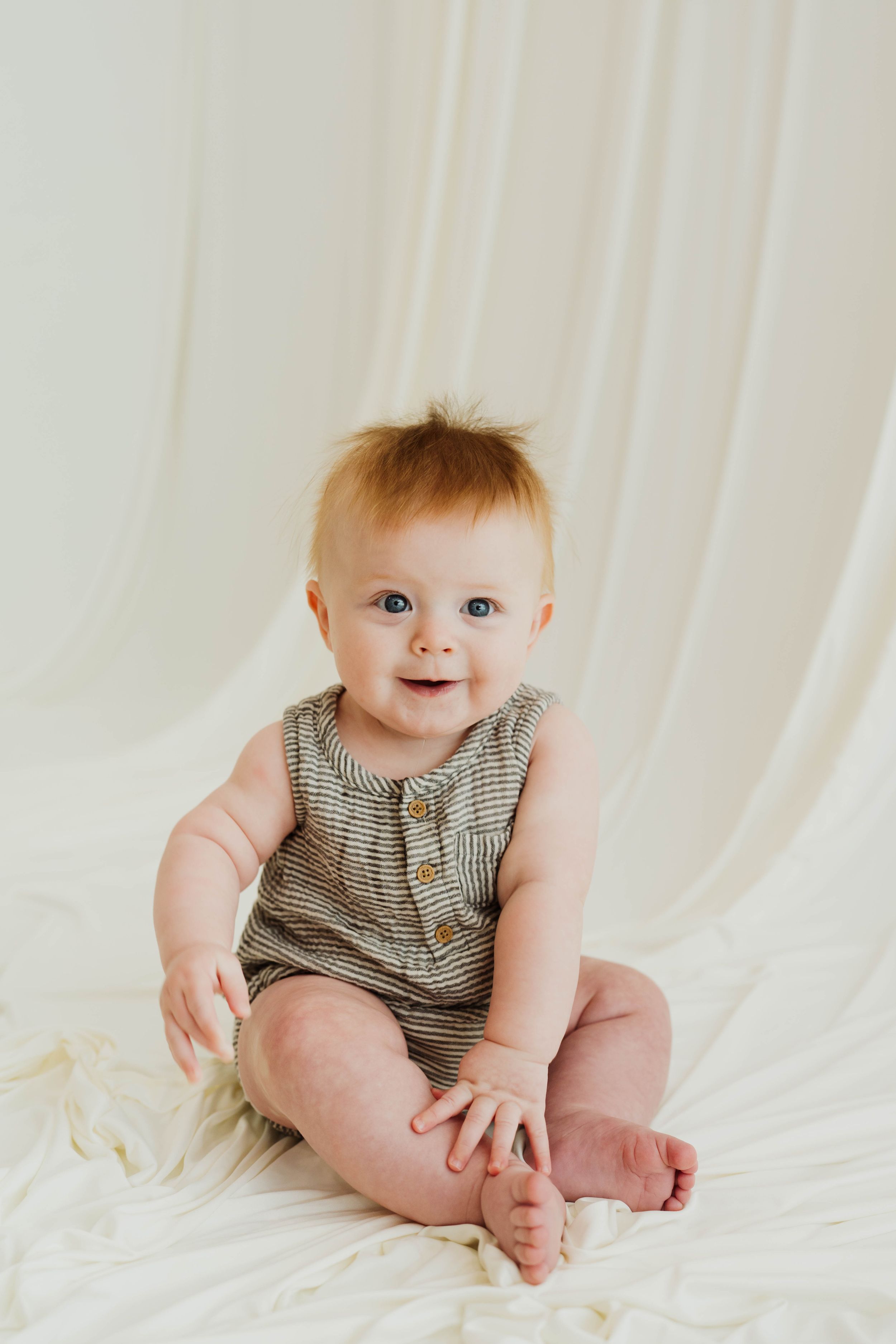 Smiling baby with blue eyes wearing a striped romper sits on white fabric backdrop.