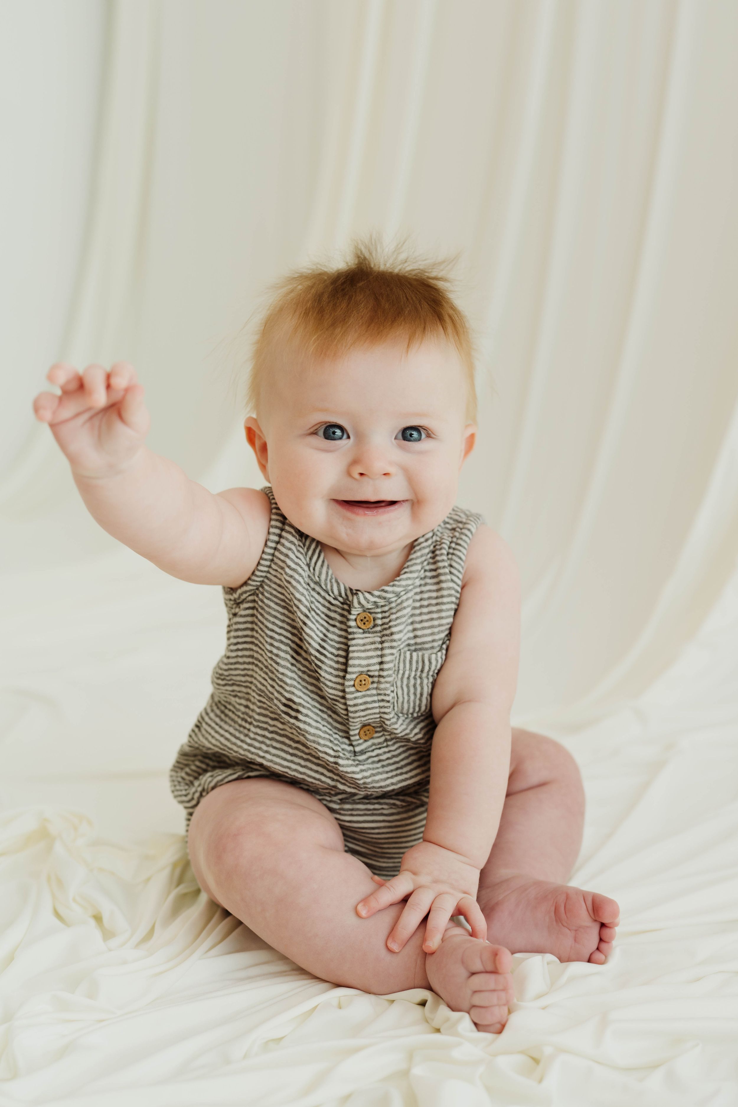 Smiling baby in striped romper sitting on white blanket, raising one hand, with light background.