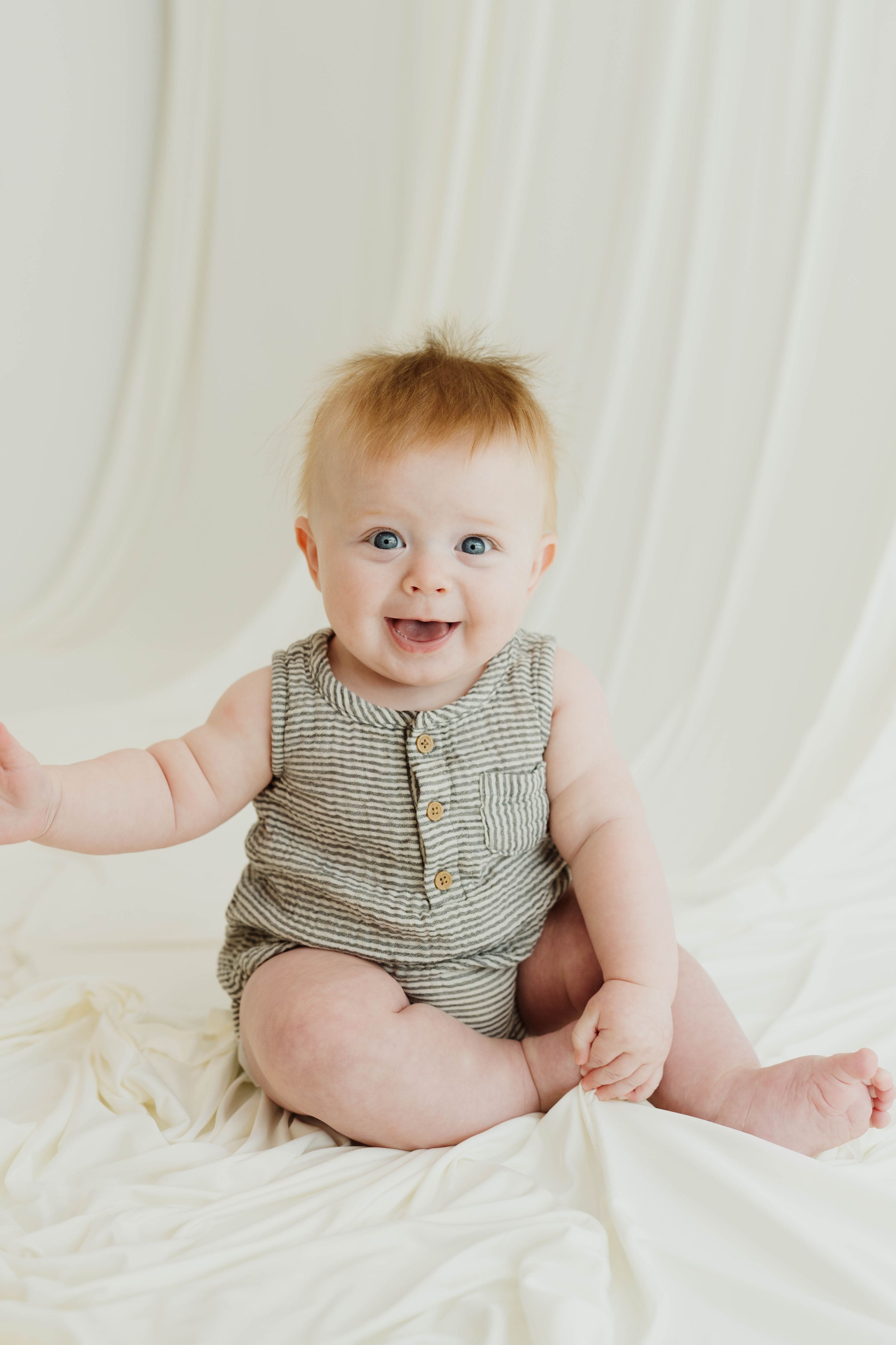 Smiling baby with blue eyes wearing a striped romper sitting on a white blanket.