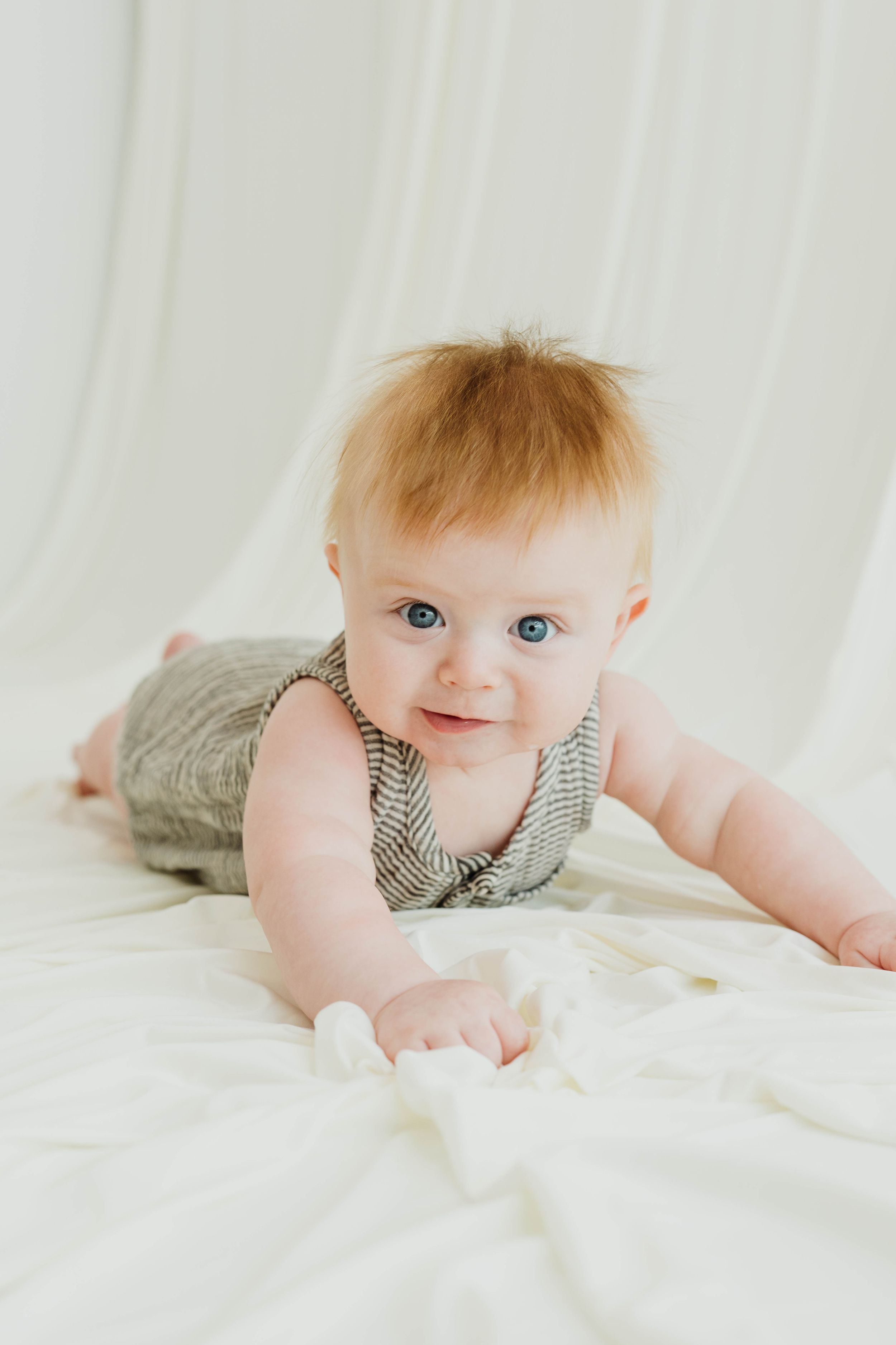 Cute red-haired baby with blue eyes doing tummy time on a white blanket in a striped romper.