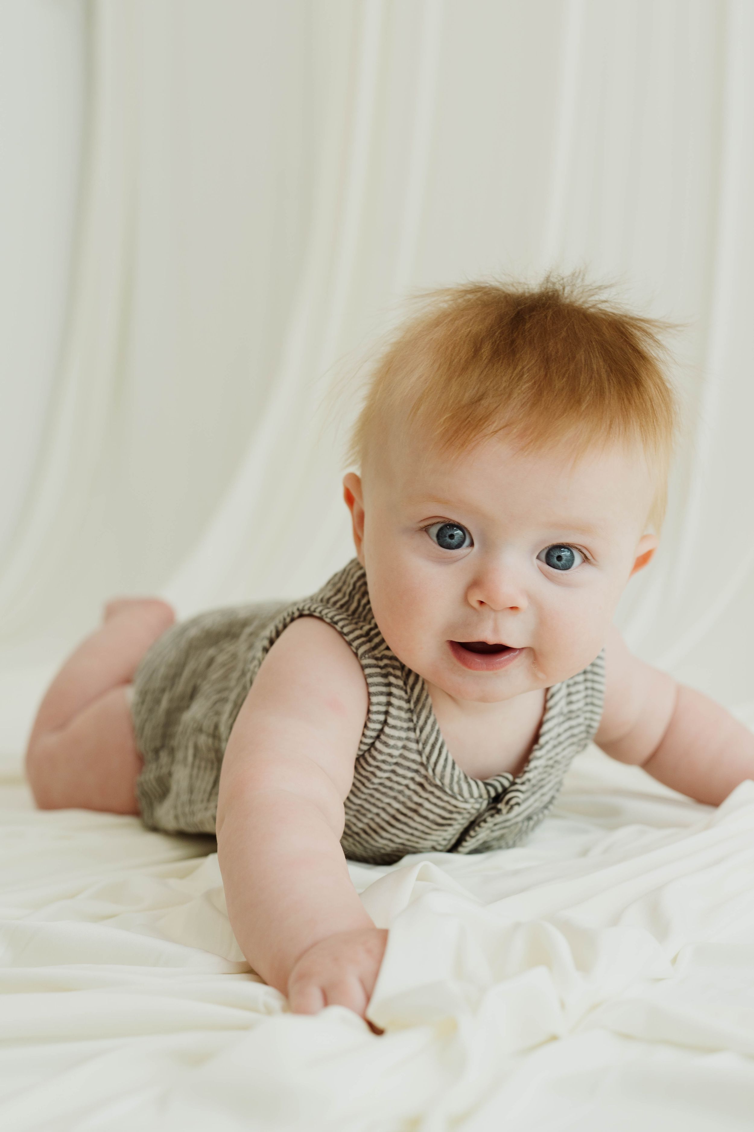 Smiling baby with blue eyes and red hair lying on white fabric wearing a gray striped romper.
