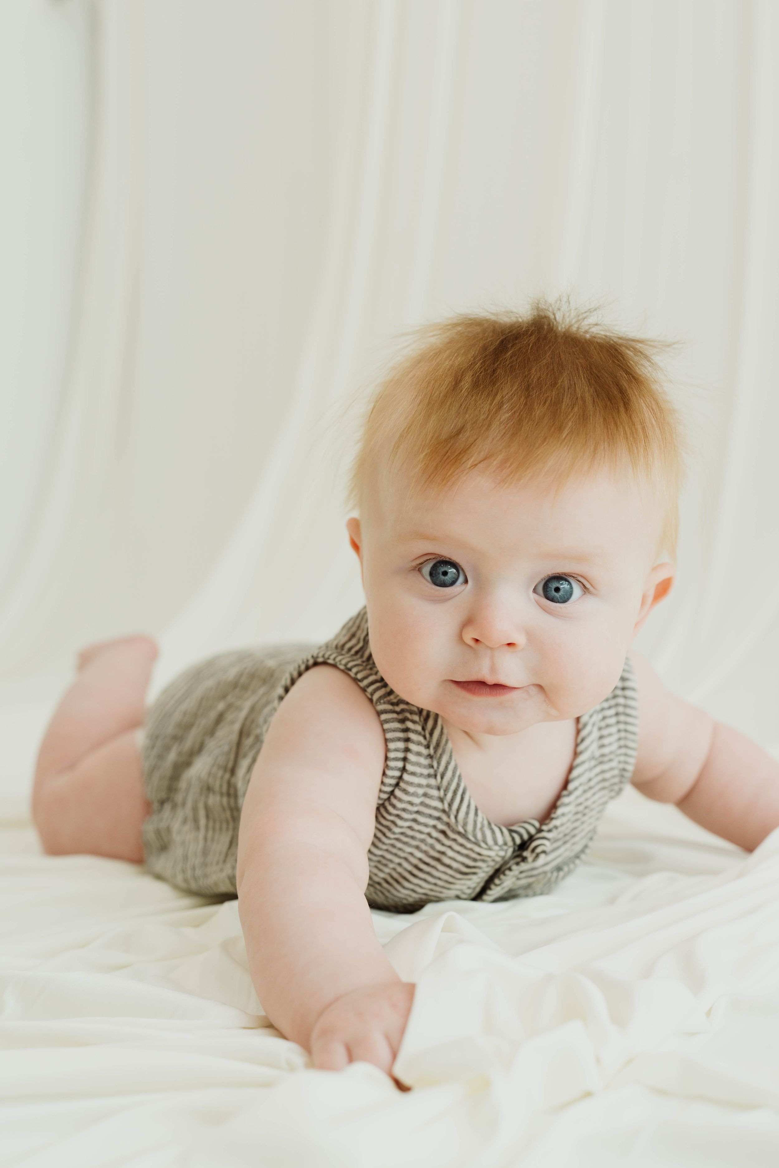 Adorable baby with blue eyes and red hair lying on white fabric wearing a striped romper.
