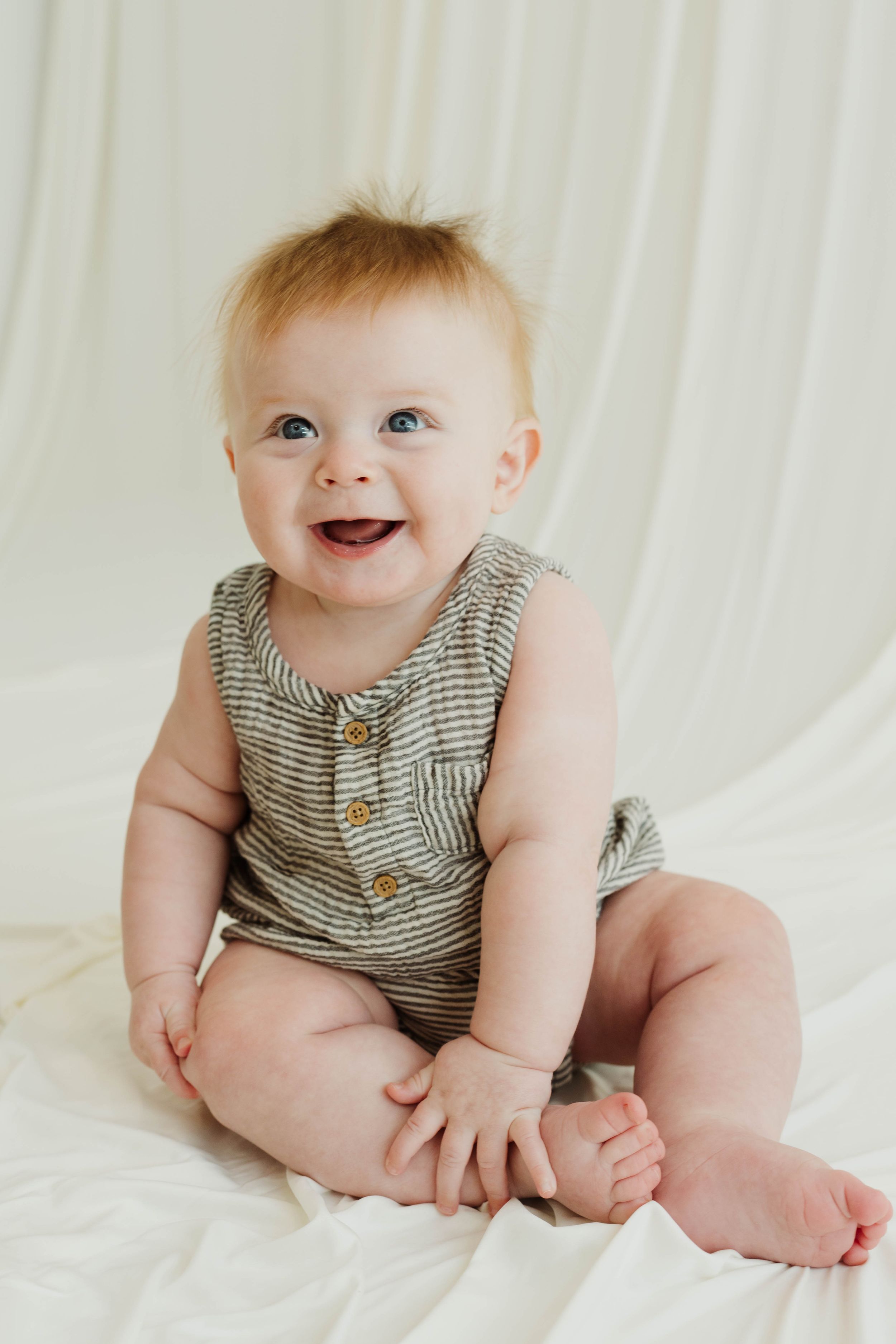 Smiling baby with blue eyes wearing a striped romper sits on a white blanket, looking happy and playful.