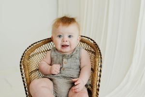 Smiling baby with blue eyes sitting in a wicker chair wearing a striped romper against a white background.