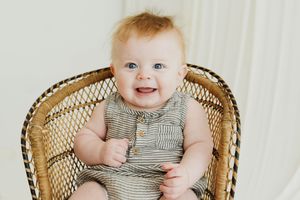 Smiling baby with blue eyes sitting in a wicker chair wearing a striped romper.