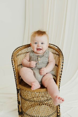 Smiling baby in striped romper sitting in a wicker chair against a white backdrop.