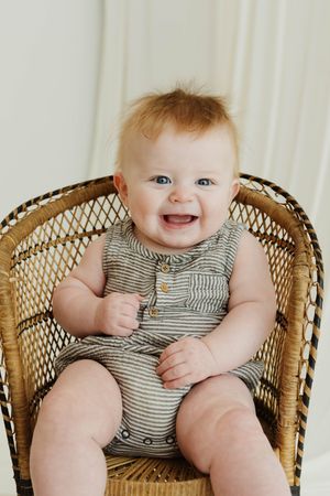 Happy baby with blue eyes in a striped romper sitting in a wicker chair.