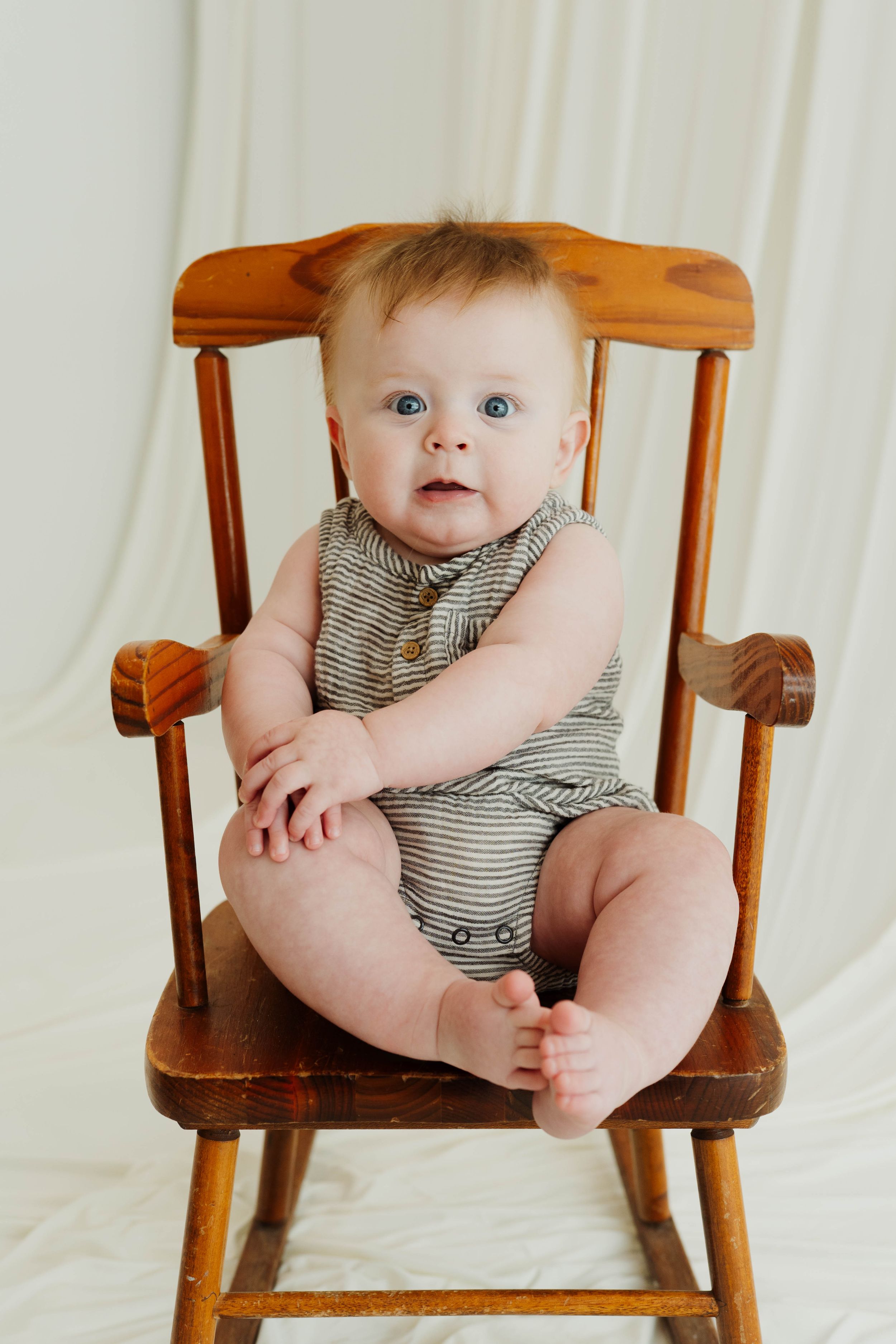 Adorable blue-eyed baby sitting in a wooden rocking chair wearing a gray striped sleeveless romper.