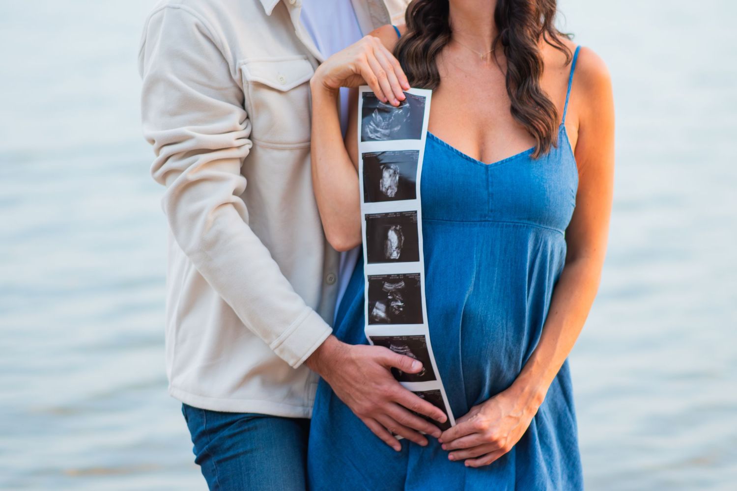 Pregnant couple holding ultrasound photos near water, woman in blue dress with partner's hands on baby bump.