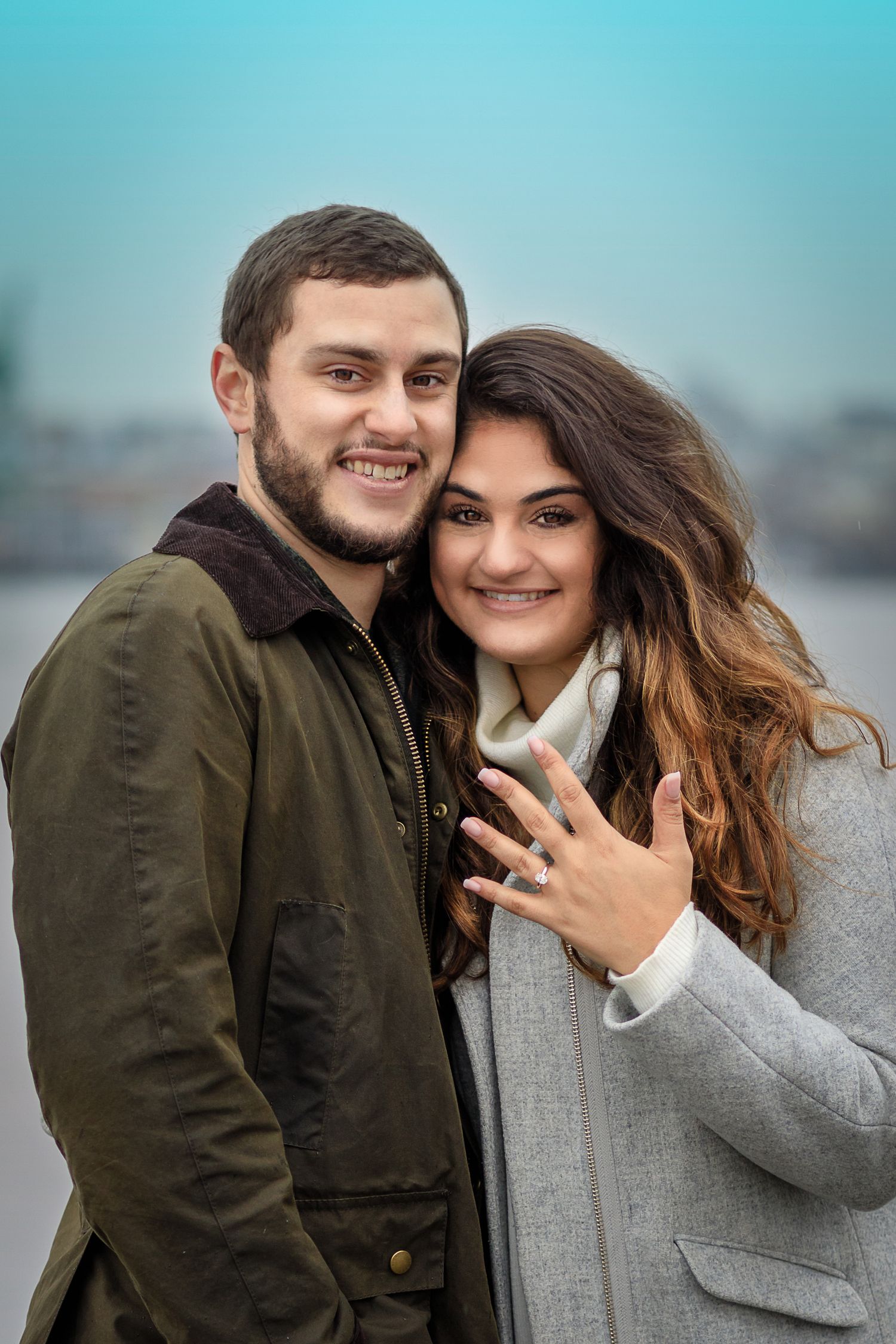 Happy couple smiling outdoors, woman showing engagement ring on hand, man in green jacket, city background.