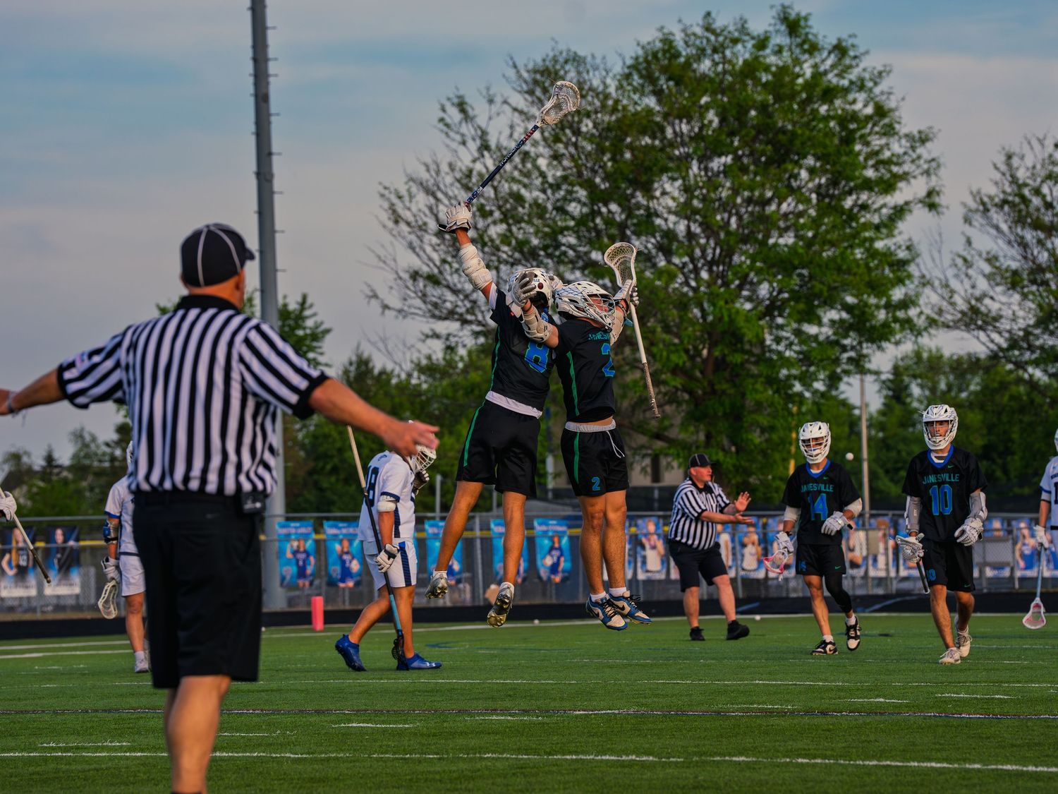 Two Janesville lacrosse players leap and bump chests to celebrate a goal as a referee signals and teammates run in to join.