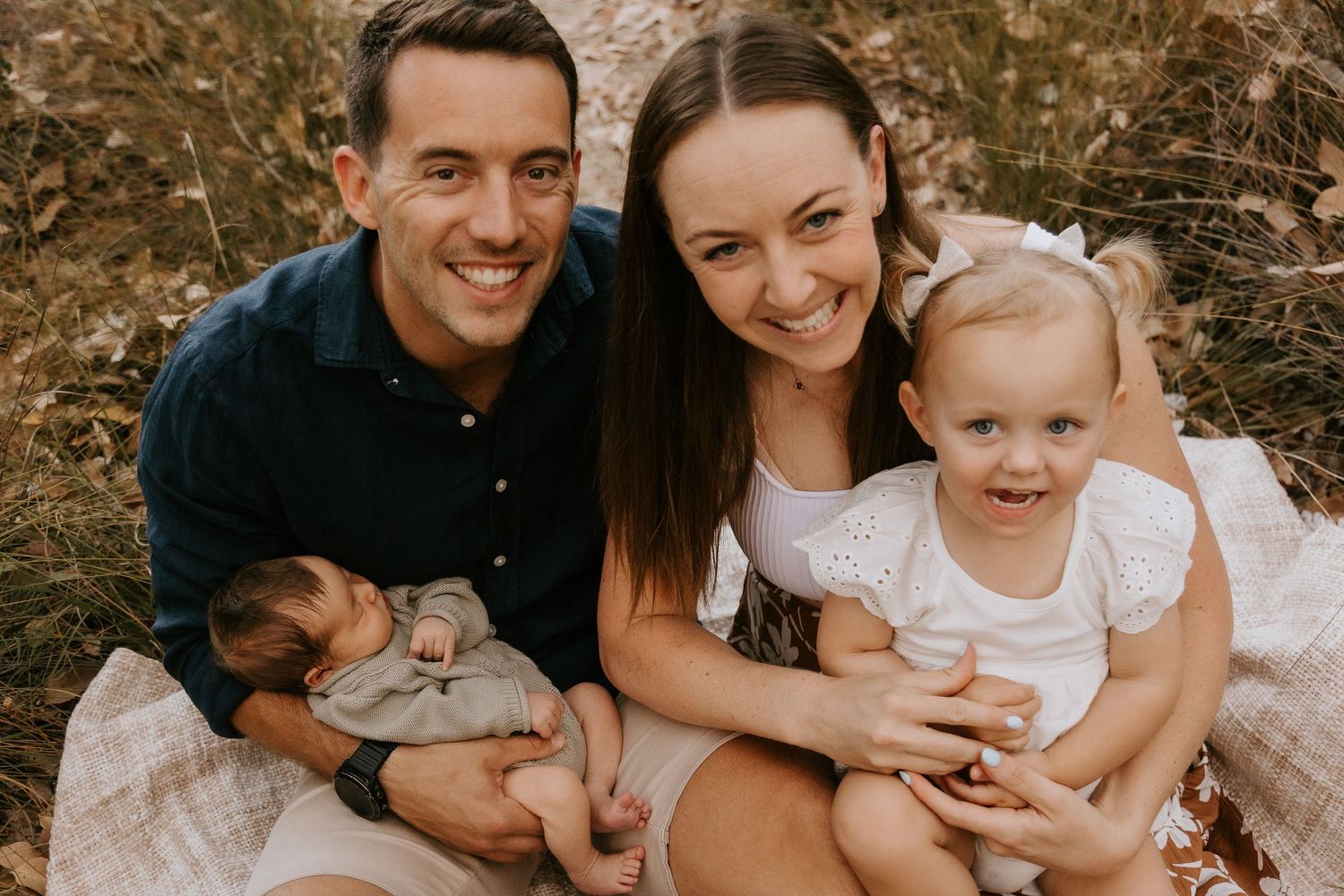 Happy family of four posing outdoors, parents smiling with a toddler girl and newborn baby on a picnic blanket.
