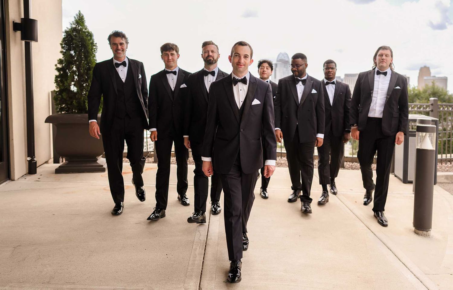 Groom leads groomsmen in black tuxedos walking confidently outdoors at a wedding venue.