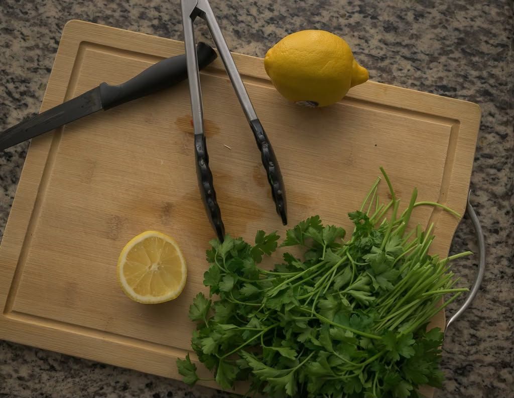Wooden cutting board with fresh cilantro, tongs, whole lemon, and halved lemon on granite countertop.