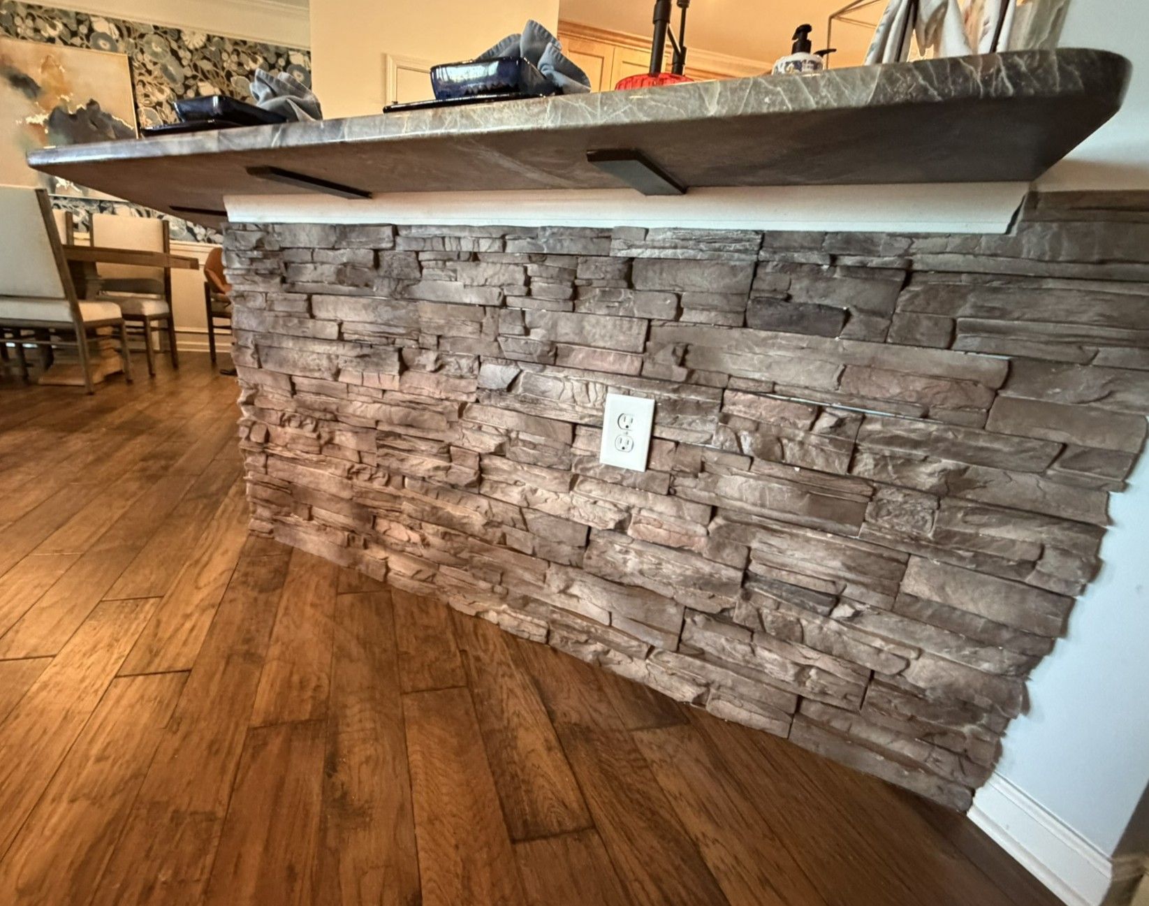 Kitchen island wrapped in rustic stacked stone veneer panels with dark granite countertop on hardwood floor.