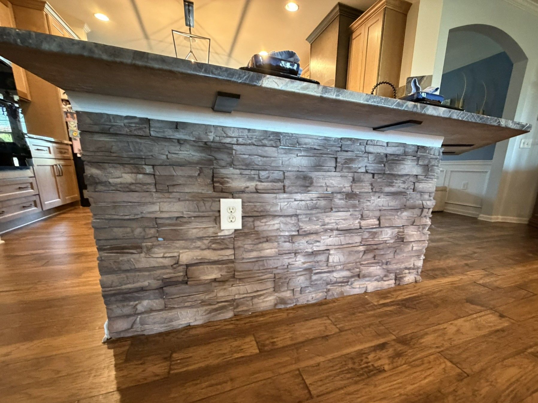 Stone veneer kitchen island with gray stacked stone panels and concrete countertop over hardwood floors.