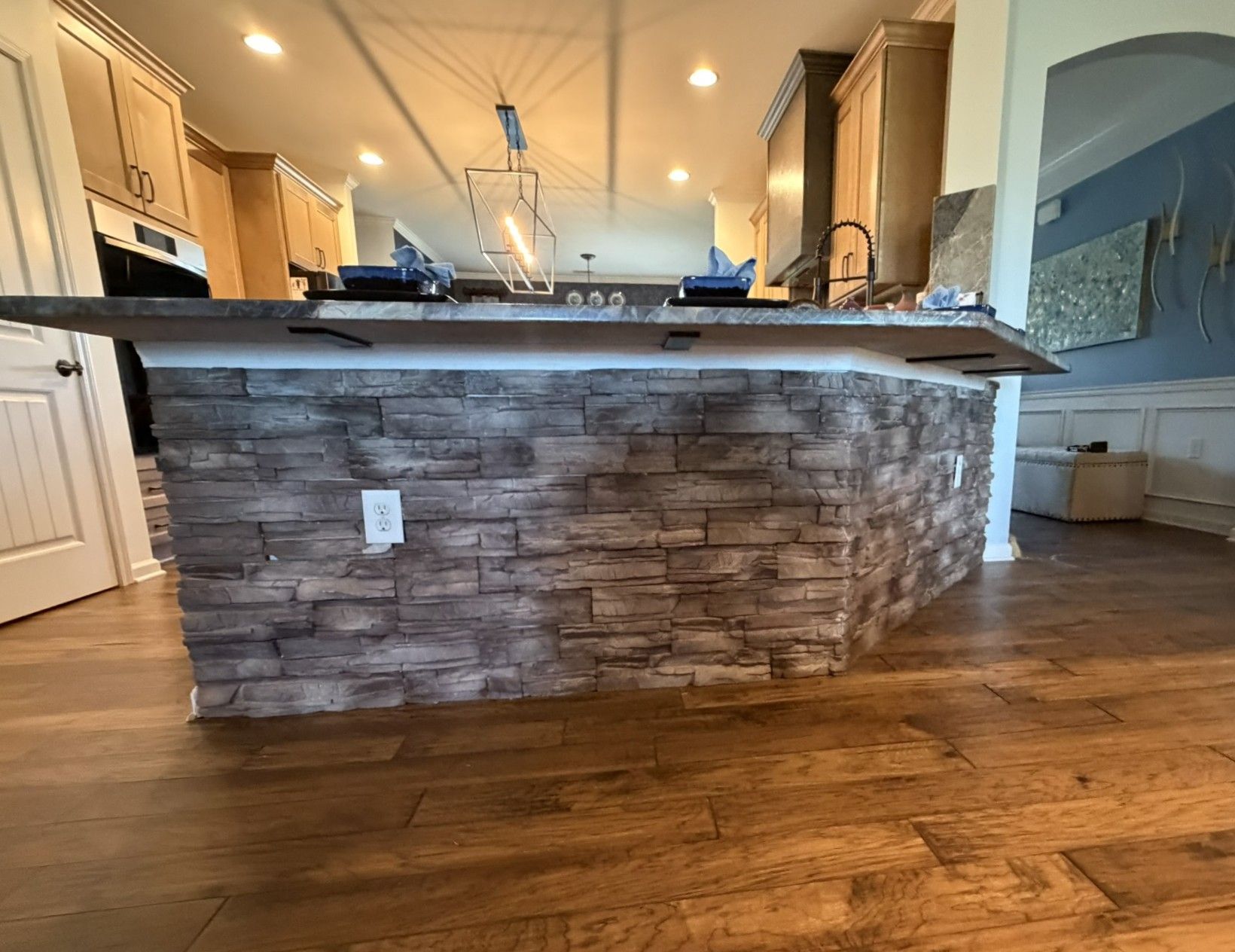 Large kitchen island clad in gray-brown stacked stone veneer with dark countertop in open-concept kitchen.