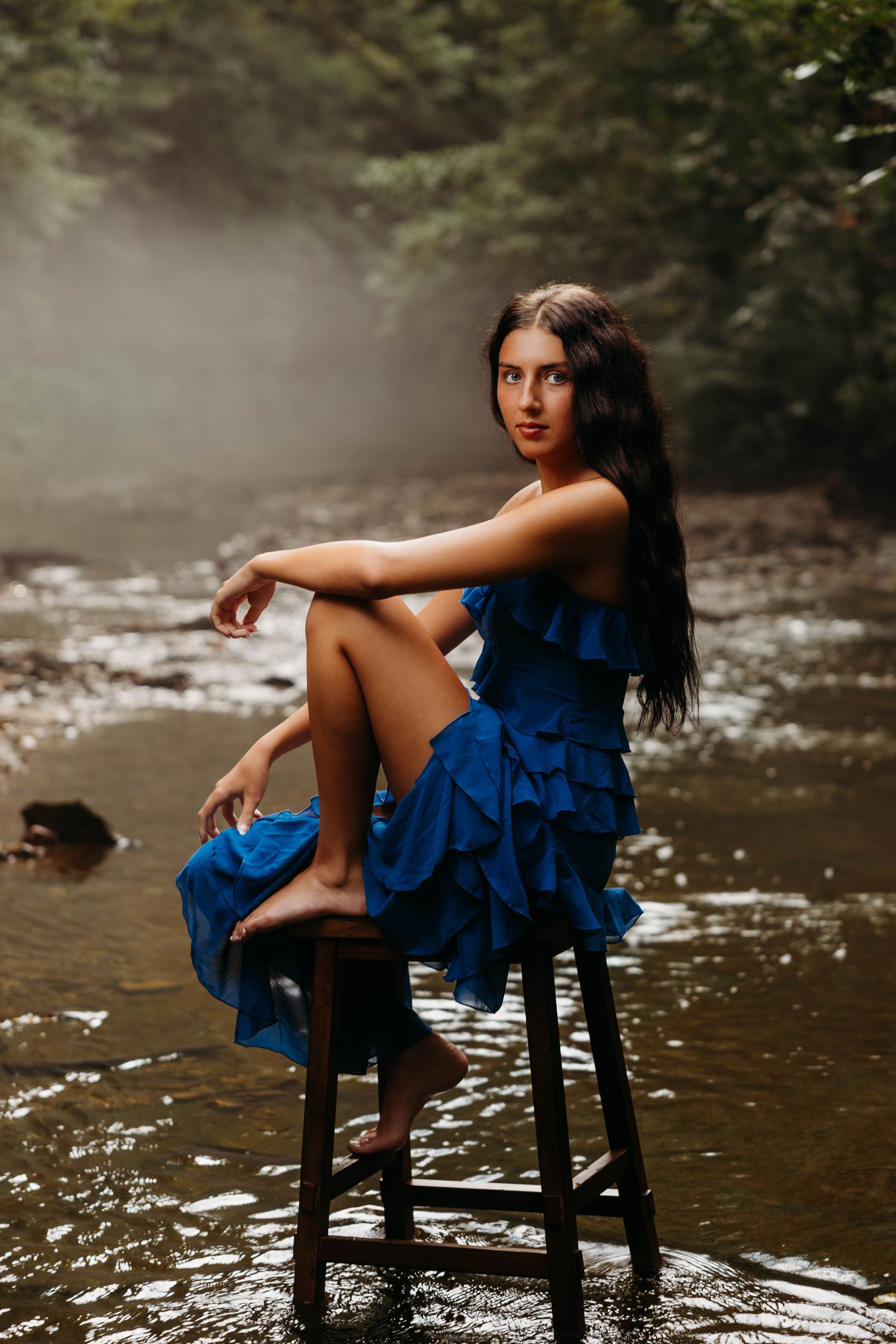 A woman in a blue ruffled dress sits barefoot on a stool in a misty forest stream.