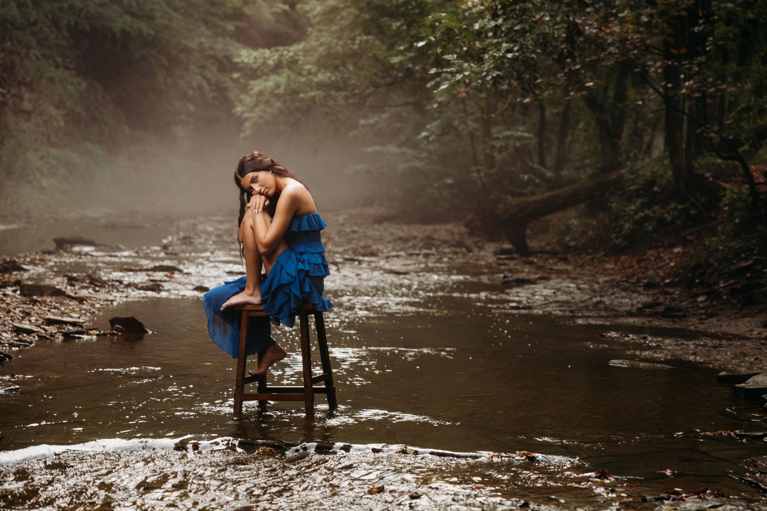 Woman in blue dress sitting on stool in misty forest river, surrounded by rocks and trees.