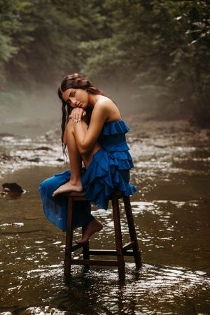A woman in a blue ruffled dress sits barefoot on a wooden stool in a misty forest stream.