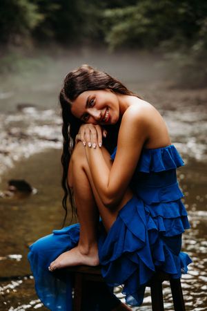 A young woman in a blue ruffled dress sits barefoot by a misty creek, smiling warmly with her knees drawn up.