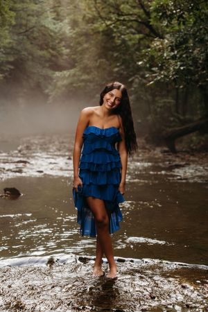 Woman in blue ruffled strapless dress standing barefoot in a misty forest creek, smiling joyfully.