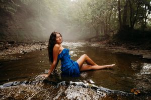 Woman in blue dress sitting on rocks in a misty forest stream, surrounded by lush green trees.
