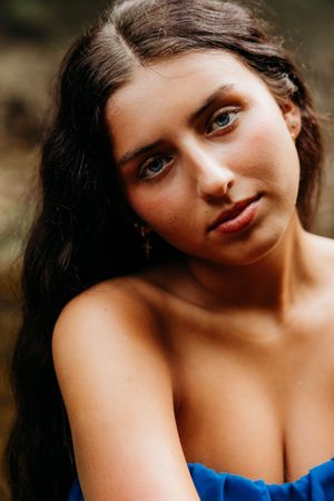 Young woman with dark wavy hair and blue eyes wearing a blue strapless dress, posing outdoors.