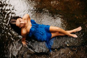 Woman in blue dress lying gracefully in a shallow rocky stream, eyes closed, water flowing around her.