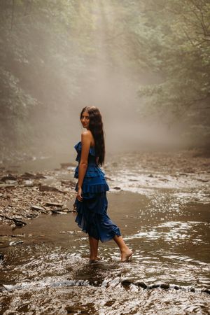 Young woman in blue ruffled dress wading barefoot in a rocky stream surrounded by misty forest trees.