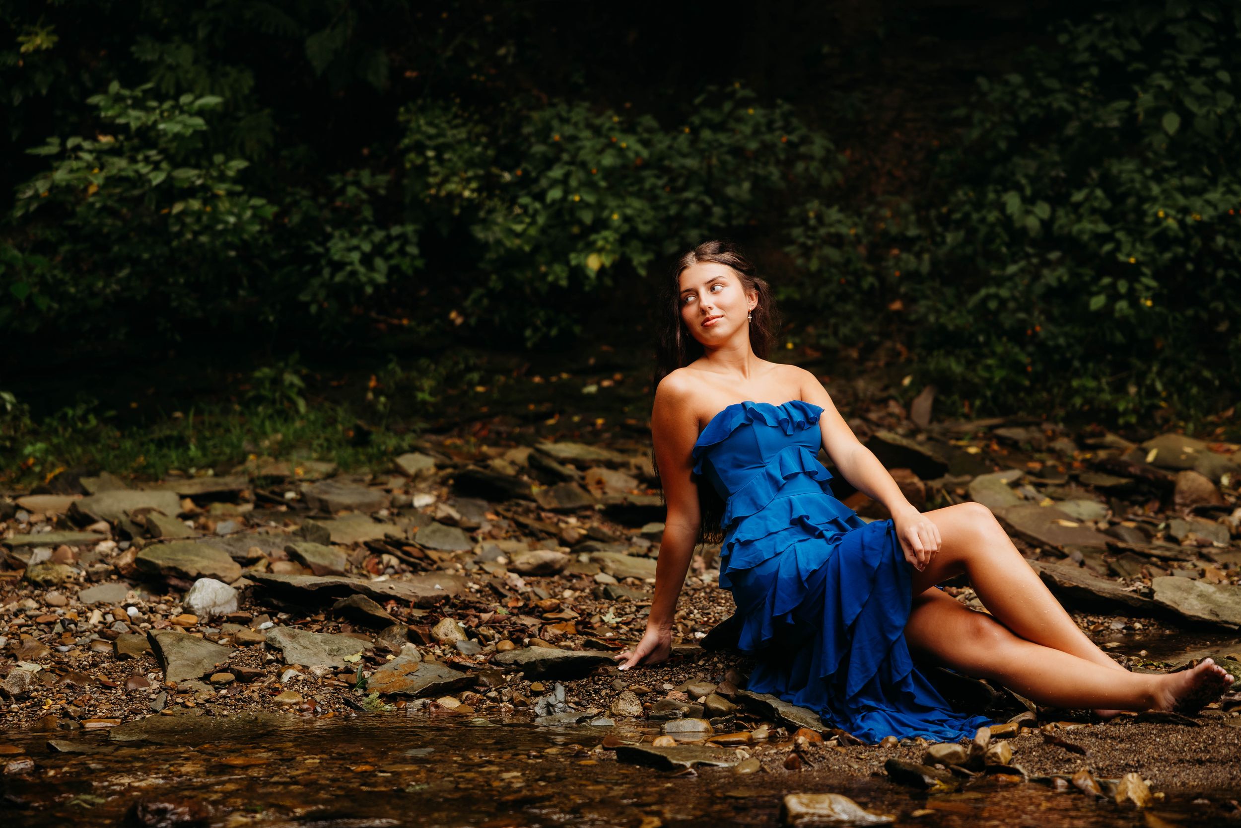 Woman in blue strapless dress sitting on rocky riverbank surrounded by lush green forest.