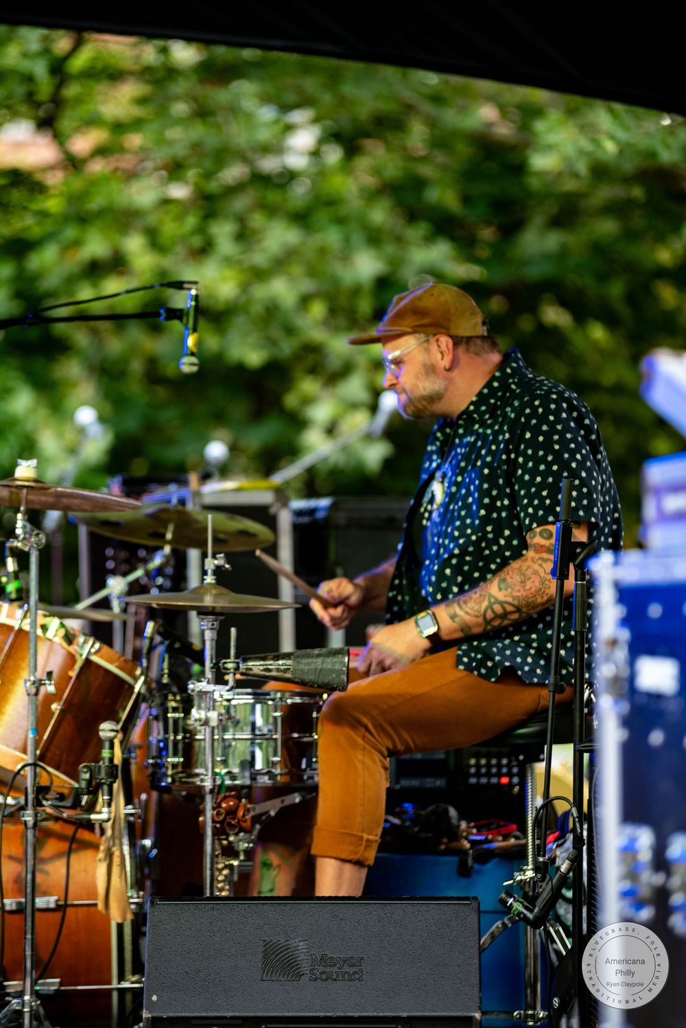 Tattooed drummer in polka dot shirt and brown cap performs on stage at an outdoor music festival.