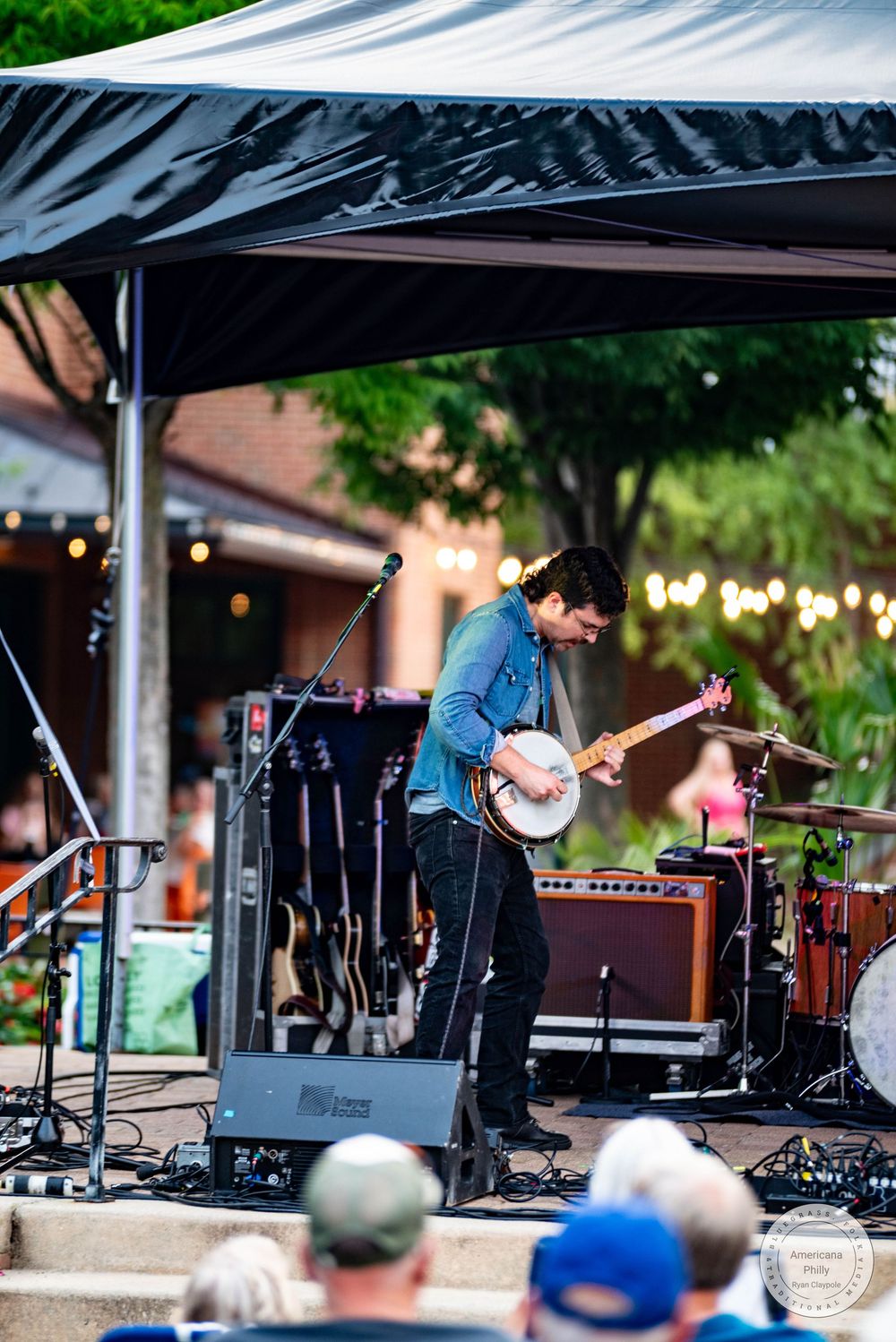 Musician playing banjo on outdoor stage under black canopy at evening concert with string lights in background.