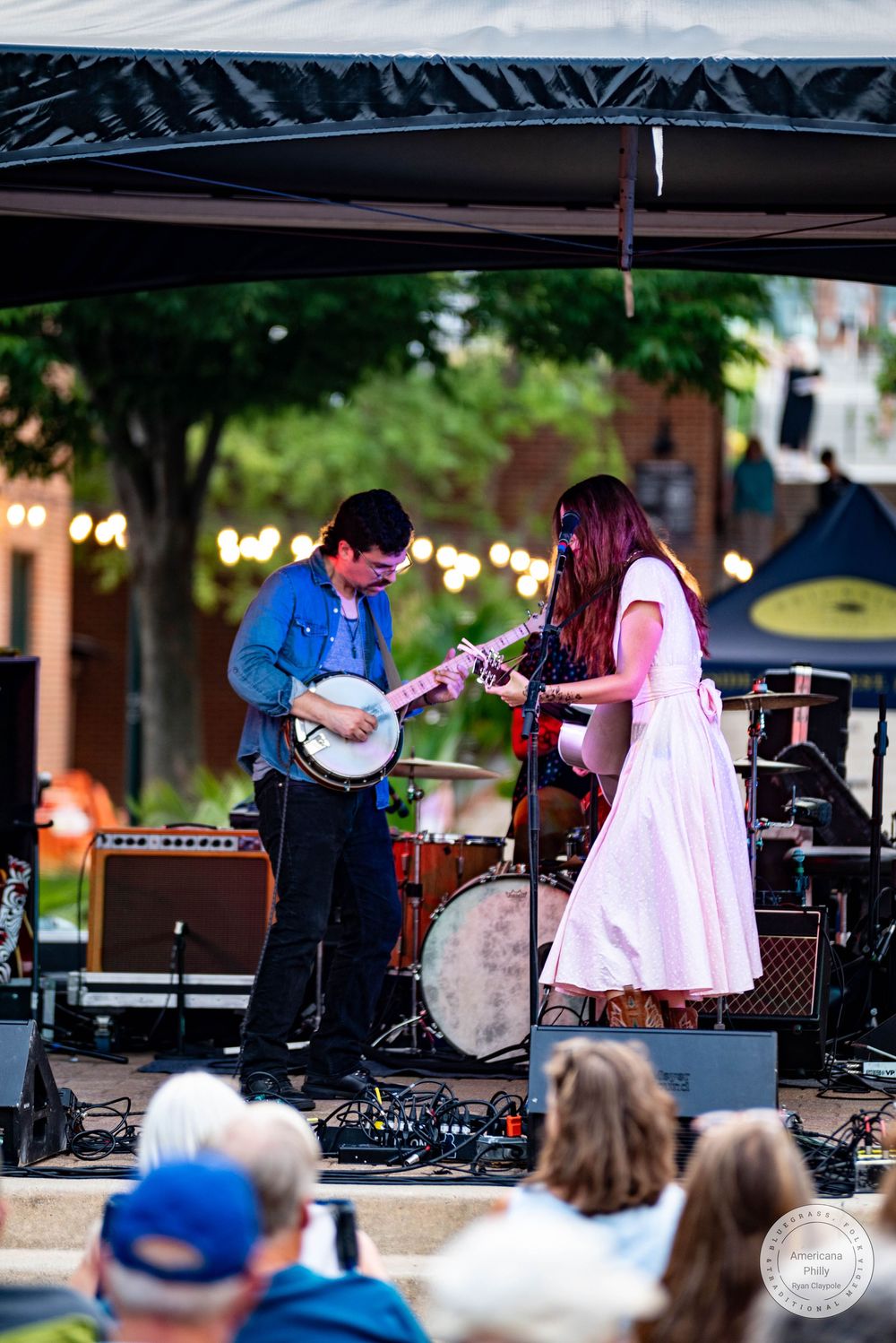 Two musicians perform on an outdoor stage, one playing banjo and one playing guitar, under string lights at a live music event.