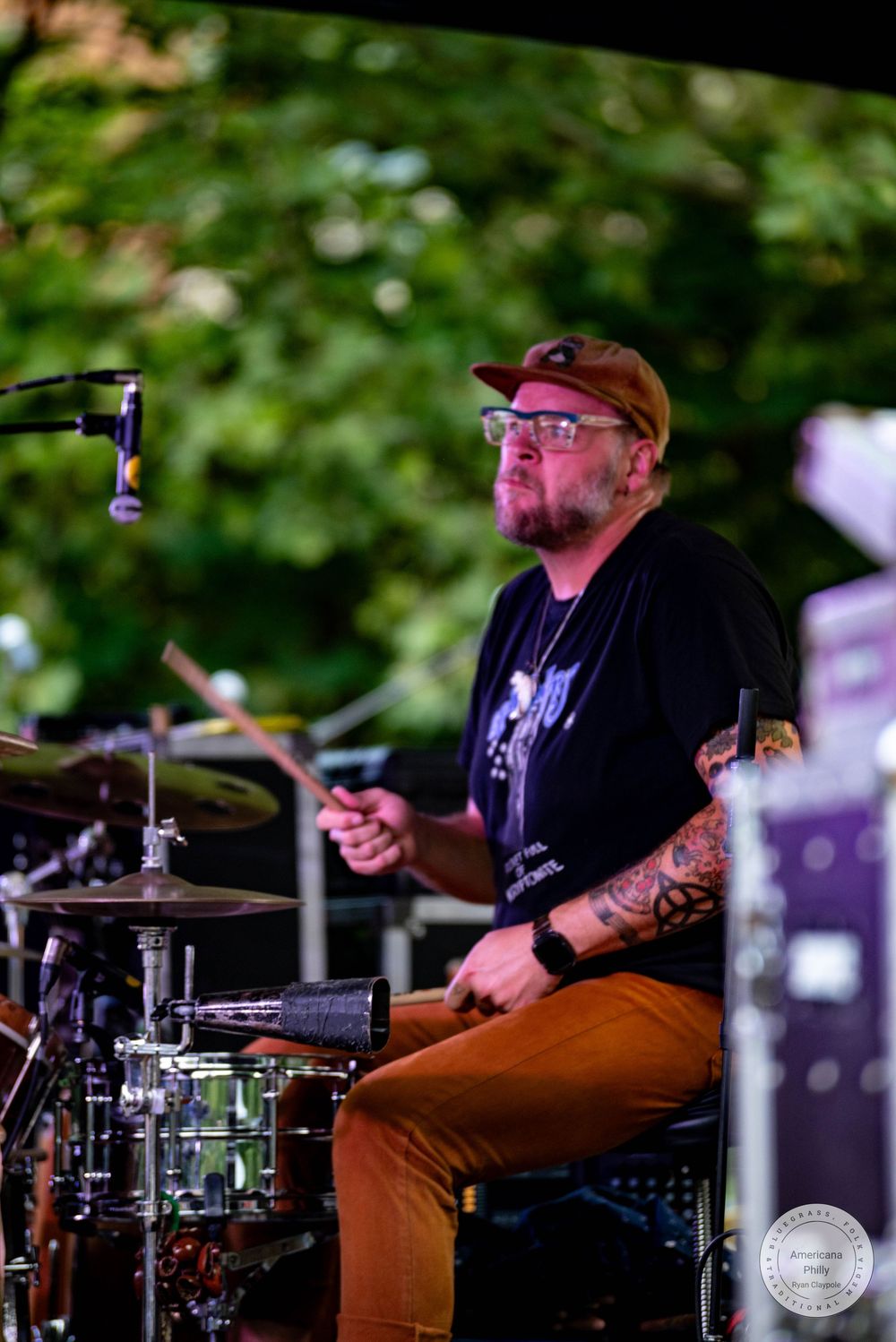 Tattooed drummer with glasses and cap plays cymbals on outdoor stage surrounded by green trees.
