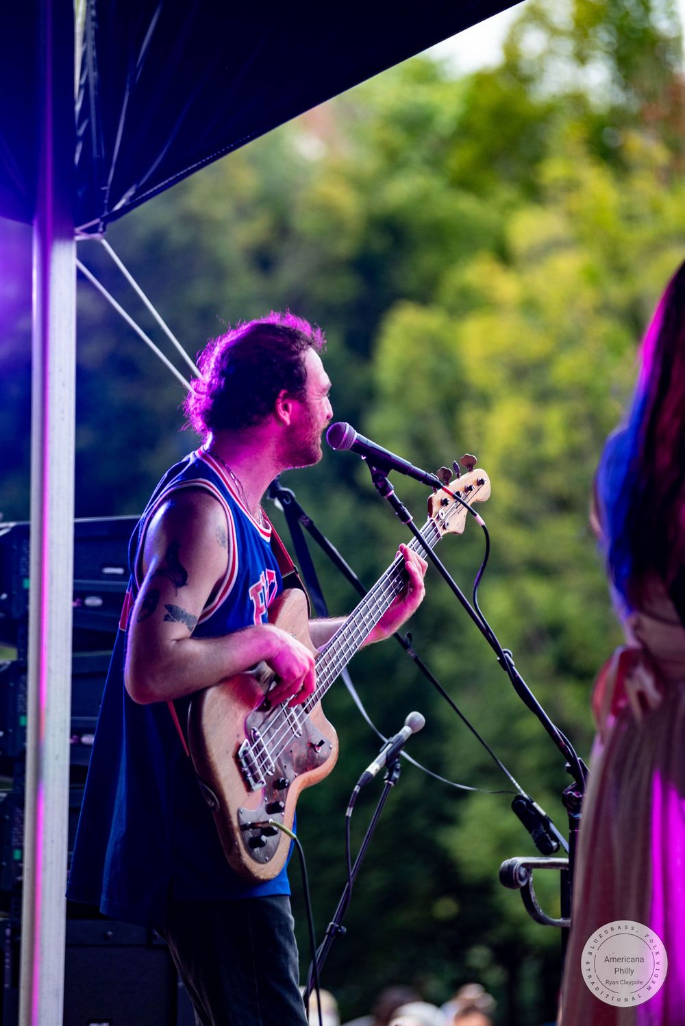 Male guitarist singing into microphone on outdoor stage under purple lights at live music festival.
