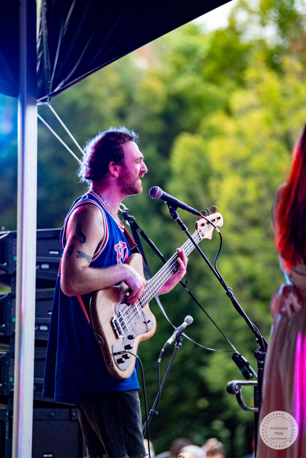 Male guitarist singing into microphone on outdoor stage, wearing blue tank top, performing at summer music festival.