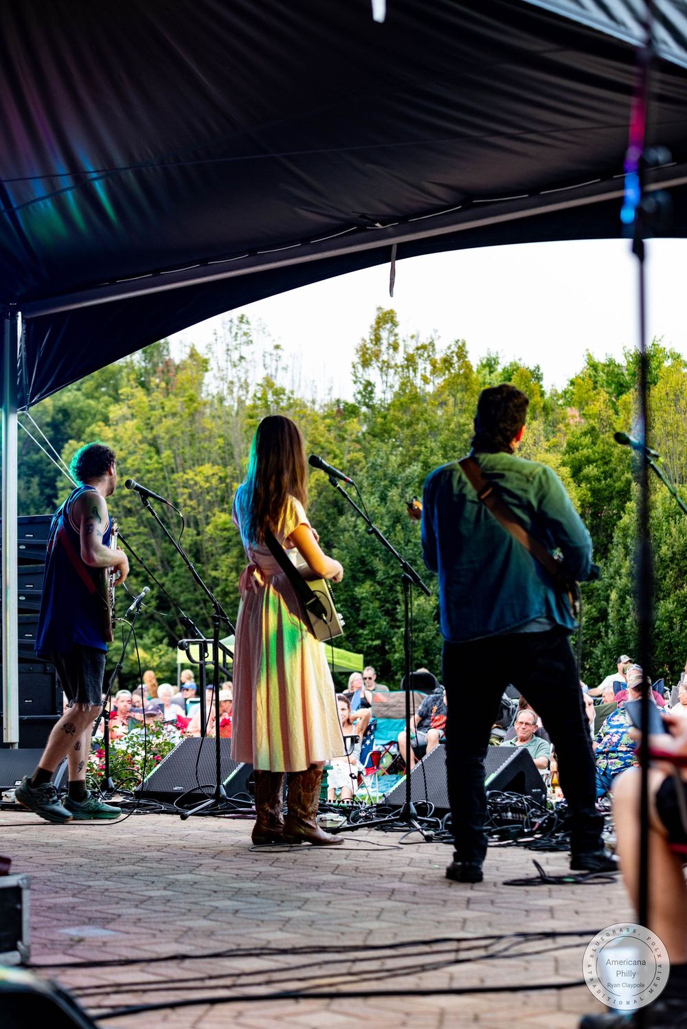 Three musicians perform on an outdoor stage at a festival, with a colorful crowd and green trees in the background.